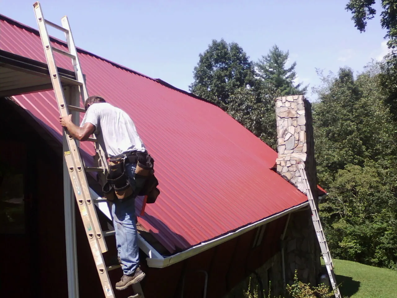 Roofer on a ladder beside a red metal roof and stone chimney — Soffit & Fascia Repair by Roofing Brothers in Long Beach